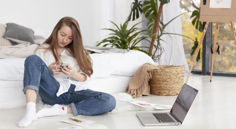 Junge Frau mit Smartphone und Notebook sitzt auf dem dem Boden vor Ihrem Bett